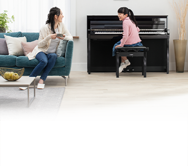 Mother enjoying tea on sofa while daughter smiles at piano in a cozy, modern living room.