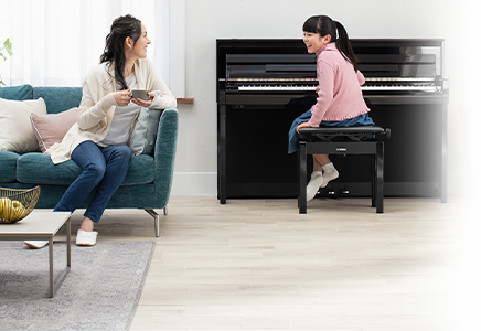 Woman enjoying coffee on couch, smiling at child playing piano in modern living room.