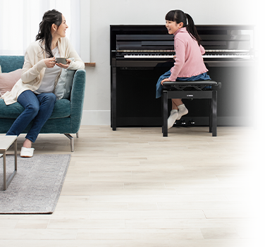 Woman drinking coffee while girl plays piano in a bright living room setting.